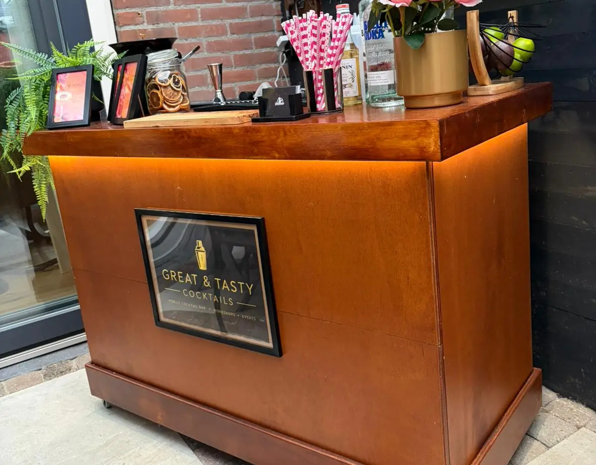 De houten mobiele cocktailbar van Great & Tasty op een terras, met het logo achter glas aan de voorkant en verlichting onder het werkblad
