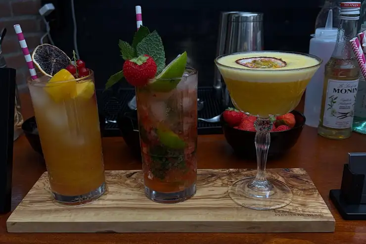 Drie cocktails op een olijfhouten plank, met shakers en flessen erachter op de bar